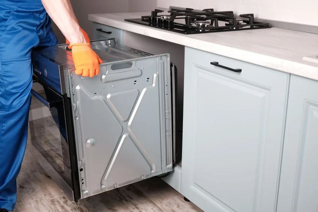 Ein Küchenmonteur in blauer Arbeitskleidung und orangefarbenen Handschuhen schiebt einen Einbau-Backofen unter ein Kochfeld in die Küchenzeile ein.