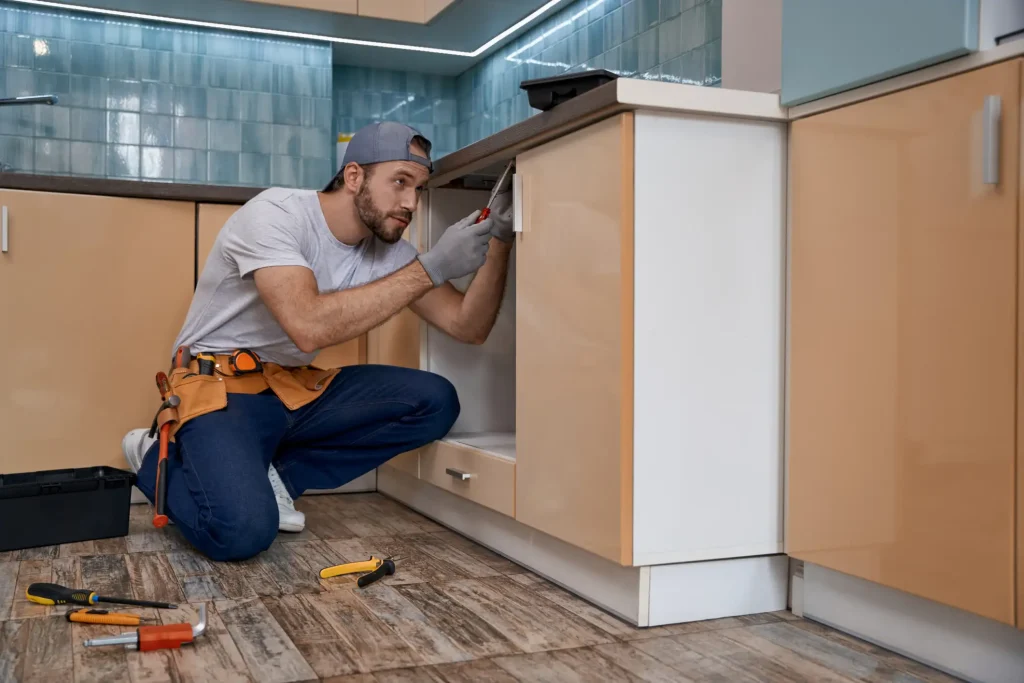 Ein Handwerker kniet in einer modernen Küche und montiert mit einem Schraubendreher ein Türscharnier an einem Unterschrank im Rahmen einer Küchenmontage.