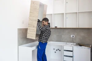 Ein Küchenmonteur hebt einen Oberschrank in Holzoptik an, um ihn an der Wand über den Unterschränken bei einer Küchenmontage zu befestigen.