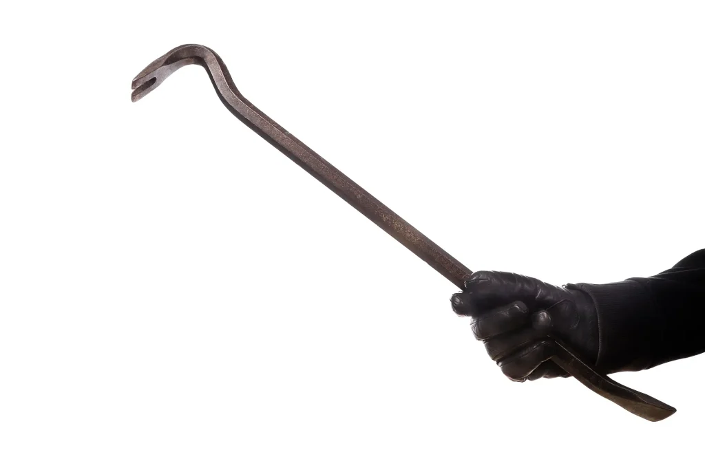 Eine Hand in einem schwarzen Lederhandschuh hält ein schmiedeeisernes Brecheisen beziehungsweise einen Kuhfuß schräg vor einem weißen Hintergrund.
