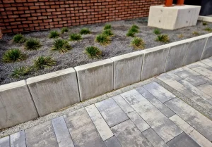 Eine Reihe gesetzter L-Steine aus Beton dient als Beetbegrenzung und Stützmauer für ein mit Ziergräsern und Split bepflanztes Beet neben einem gepflasterten Weg.