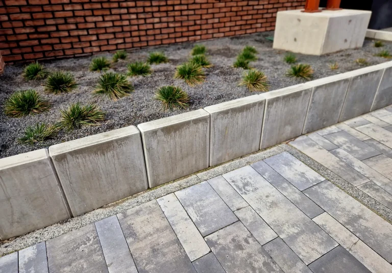 Eine Reihe gesetzter L-Steine aus Beton dient als Beetbegrenzung und Stützmauer für ein mit Ziergräsern und Split bepflanztes Beet neben einem gepflasterten Weg.