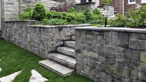 Eine terrassierte Natursteinmauer aus grau-braunen Bruchsteinen mit integrierter Steintreppe, die zu einem bepflanzten Hochbeet und Vorgarten führt.
