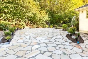 Große, geschwungene Gartenterrasse aus hellen Naturstein-Polygonalplatten mit breiten Fugen. Die neu angelegte Steinterrasse schließt an ein Wohnhaus an und wird von kleinen Pflanzbeeten sowie dichten, grünen Bäumen im Hintergrund umgeben.