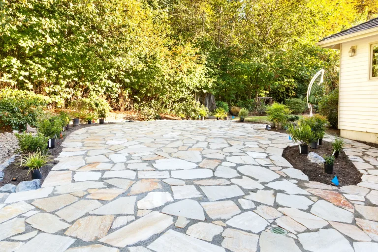 Große, geschwungene Gartenterrasse aus hellen Naturstein-Polygonalplatten mit breiten Fugen. Die neu angelegte Steinterrasse schließt an ein Wohnhaus an und wird von kleinen Pflanzbeeten sowie dichten, grünen Bäumen im Hintergrund umgeben.