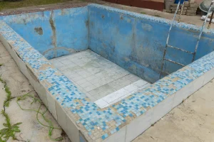 Blick in das Innere eines leeren, sanierungsbedürftigen Pools. Die Wände sind in verwittertem Hellblau gestrichen, während der Betonboden starke Verschmutzungen, dunkle Flecken und etwas Laub aufweist. Auf der rechten Seite führt eine gemauerte Treppe nach oben, daneben befinden sich alte Pool-Düsen und Anschlüsse in der Wand.