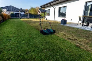 Ein elektrischer Vertikutierer steht auf einer Rasenfläche hinter einem modernen Wohnhaus. Die linke Seite zeigt dichten, grünen Rasen, während rechts die gelockerten Moos- und Filzreste nach dem Vertikutieren des Bodens sichtbar sind.