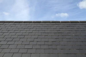 Nahaufnahme eines eingedeckten Schieferdachs mit dunklen Schieferplatten und Dachfirst vor einem leicht bewölkten blauen Himmel.