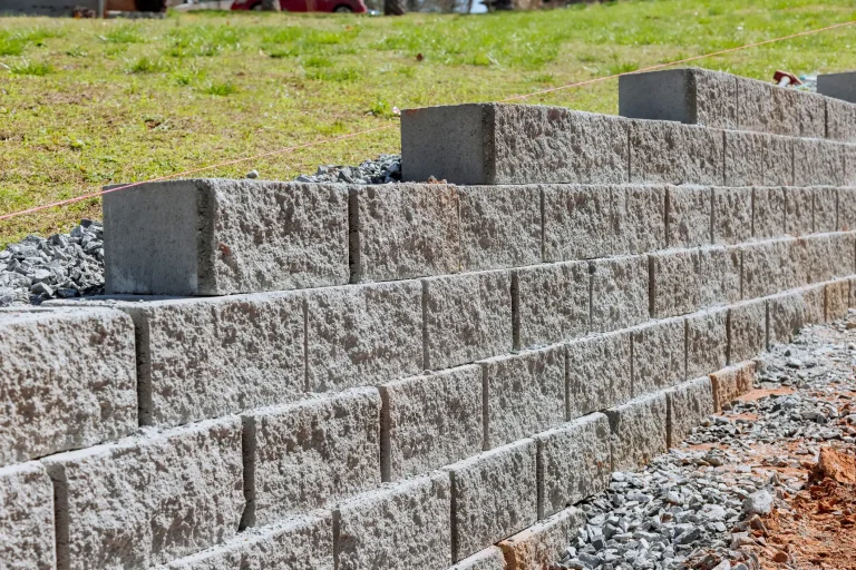 Eine im Bau befindliche Stützmauer aus grauem Beton mit strukturierter Oberfläche, die stufenförmig an einem leicht abfallenden Grashang errichtet wird.