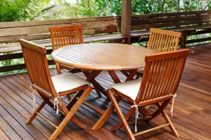 Überdachte Holzterrasse aus warmen Teak-Terrassendielen mit einer passenden Sitzgruppe aus einem runden Teakholztisch und vier Klappstühlen mit hellen Sitzkissen.
