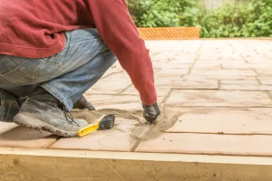 Nahaufnahme eines Gartenbauers in Jeans, roten Pullover und Arbeitsschuhen, die auf einer Terrasse kniet. Mit Handschuhen wird Fugensand präzise in die Fugen zwischen beigefarbenen Terrassenplatten eingearbeitet. Neben dem Fuß liegt eine kleine gelbe Bürste zum Einkehren des Sands auf dem Boden.