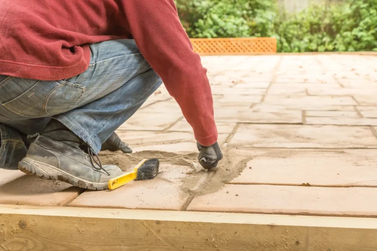 Nahaufnahme eines Gartenbauers in Jeans, roten Pullover und Arbeitsschuhen, die auf einer Terrasse kniet. Mit Handschuhen wird Fugensand präzise in die Fugen zwischen beigefarbenen Terrassenplatten eingearbeitet. Neben dem Fuß liegt eine kleine gelbe Bürste zum Einkehren des Sands auf dem Boden.