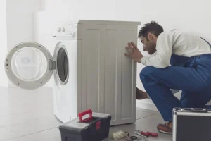 Ein Hausgeräte-Servicetechniker in blauer Latzhose kniet neben einer geöffneten weißen Waschmaschine, um sie zu überprüfen und zu reparieren.