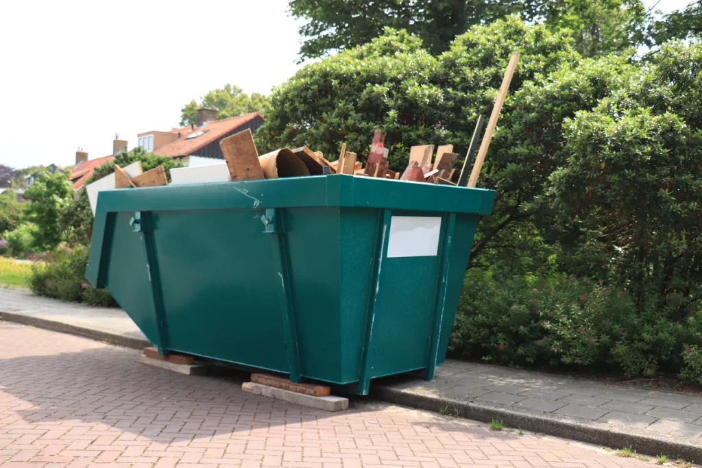 Grüner Abfallcontainer mit Holz und Holzstäben neben einem Busch.