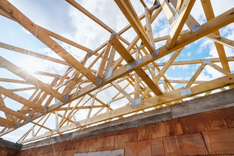 Frisch errichteter Holz-Dachstuhl aus Dachbindern und Sparren auf einer Ziegelwand im Rohbau vor sonnigem Himmel.