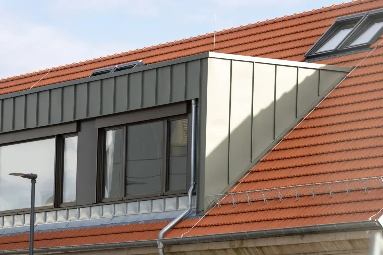 Moderne Fertiggaube mit grauer Stehfalz-Blechverkleidung und großen Fenstern auf einem roten Ziegeldach.