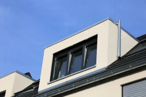 Moderne helle Flachdachgaube mit dunklem Dreifach-Fenster, Zink-Abdeckungen und Dachrinne an einem Steildach vor blauem Himmel.