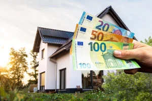 Ein modernes Einfamilienhaus mit dunklem Satteldach und grüner Gartenfläche, vor dem mehrere Euro-Banknoten in einer Hand gehalten werden.