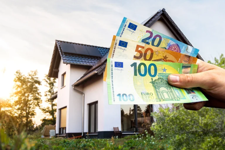 Ein modernes Einfamilienhaus mit dunklem Satteldach und grüner Gartenfläche, vor dem mehrere Euro-Banknoten in einer Hand gehalten werden.