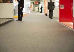 Messehalle mit grauem Teppichboden und verschwommenen Personen in Anzügen im Gang zwischen Messeständen.