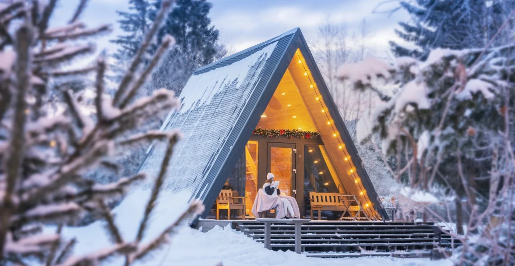 Beleuchtetes A-Frame Nurdachhaus im Winter mit Lichterkette und verschneiter Umgebung.