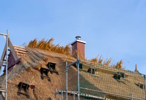 Dachdeckerarbeiten an einem Reetdach mit frischen Schilfbündeln, Gerüst und Schornstein vor blauem Himmel.