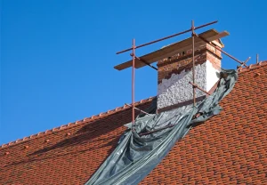 Schornsteinsanierung an einem gemauerten Kamin mit Baugerüst und Schutzfolie auf einem roten Ziegeldach vor blauem Himmel.