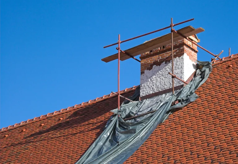 Schornsteinsanierung an einem gemauerten Kamin mit Baugerüst und Schutzfolie auf einem roten Ziegeldach vor blauem Himmel.