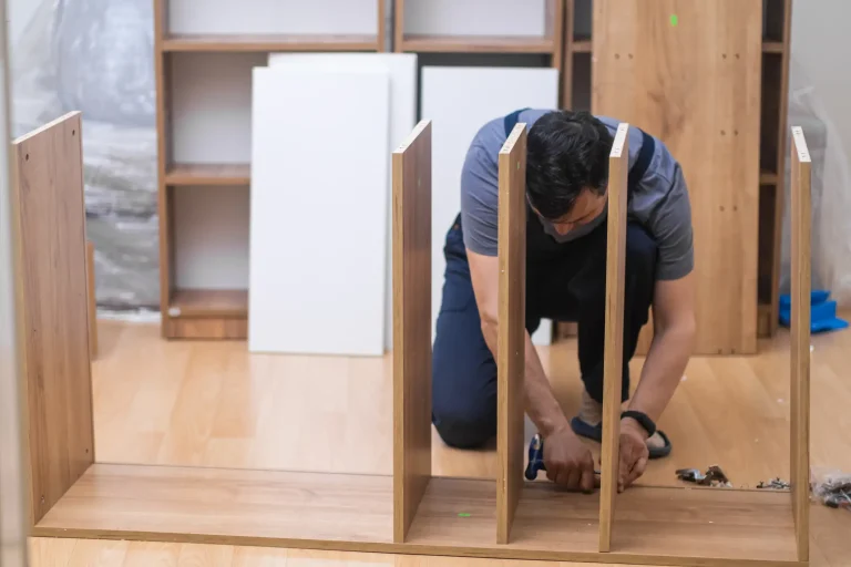 Monteur führt eine Schrankmontage durch, indem er auf dem Boden kniend Zwischenwände eines Holzregals zusammenschraubt.