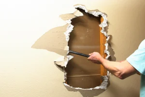 Eine Person reißt mit einem Brecheisen ein Loch in eine gelb gestrichene Trockenbauwand.