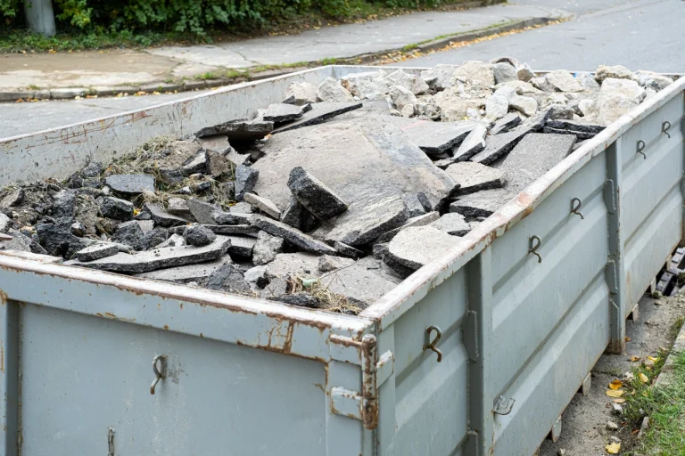 Ein grauer Absetzcontainer gefüllt mit Bauschutt, darunter Betonbrocken und dunkle Asphaltplatten, auf einem Bürgersteig.