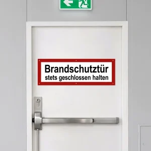 Weißes Schild mit rotem Rand auf einer Trockenbau-Brandschutztür mit der Aufschrift „Brandschutztür stets geschlossen halten“, darüber ein grünes Notausgangsschild.