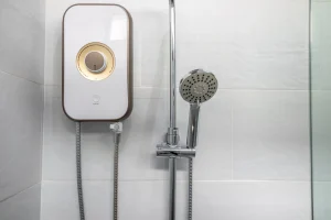 Moderner weißer Durchlauferhitzer mit goldenem Drehregler an einer gefliesten Badezimmerwand neben einer Duschbrause.