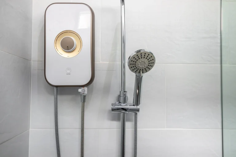 Moderner weißer Durchlauferhitzer mit goldenem Drehregler an einer gefliesten Badezimmerwand neben einer Duschbrause.