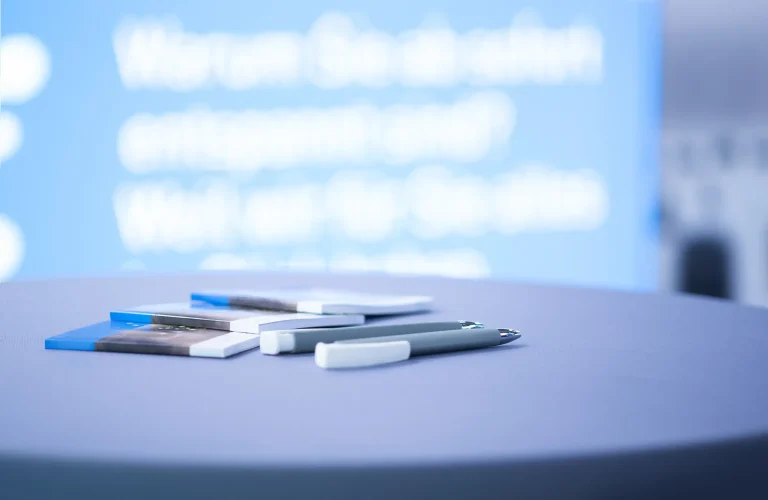 Informationsbroschüren und Stifte auf einem Stehtisch bei einer Messe mit blauem Präsentationshintergrund.