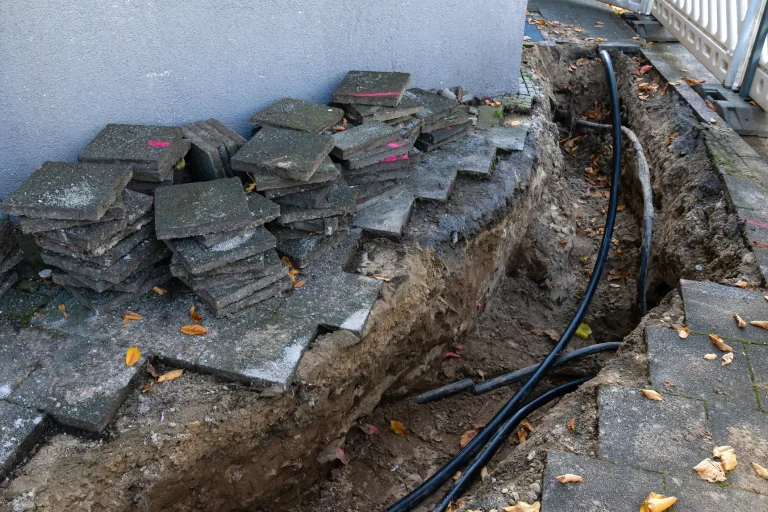 Freigelegter Kabelgraben für den Strom-Hausanschluss mit schwarzen Erdkabeln entlang einer Hauswand und aufgenommenen Gehwegplatten.