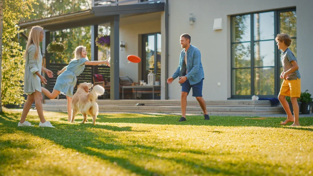 Familie spielt mit einem Hund und einer Frisbeescheibe auf dem Spielrasen im Garten.