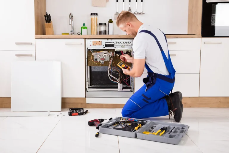 Elektriker überprüft mit einem Multimeter die elektrischen Anschlüsse einer geöffneten Spülmaschine in einer Küche.