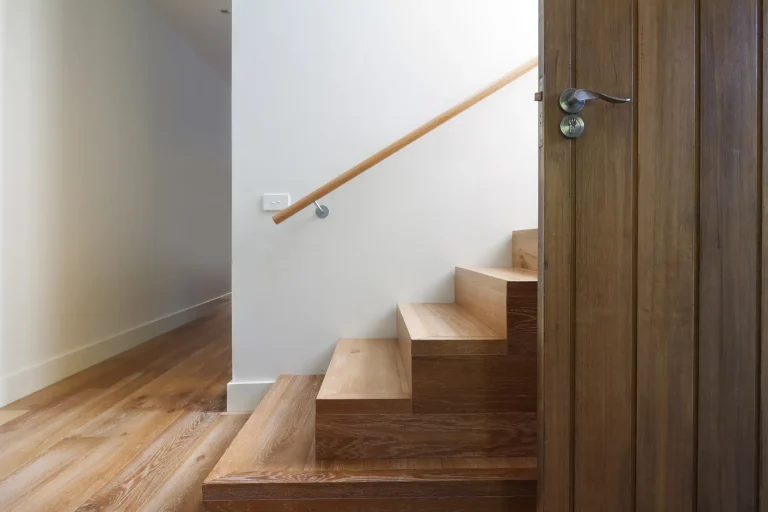 Moderne Holztreppe mit Wandhandlauf und geöffneter Eingangstür aus Holz in hellem Flur.