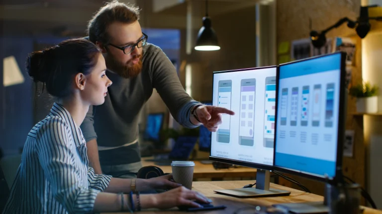 Zwei junge Kollegen arbeiten gemeinsam an einem UX/UI-Design auf einem Doppelmonitor. Der eine Kollege sitzt am Schreibtisch und bedient Maus und Tastatur, während der andere Kollege neben ihr steht und auf einen der Bildschirme zeigt, der mobile App-Layouts zeigt. Im Hintergrund ist ein ein moderne, kreativ gestaltetes Büro zu sehen.