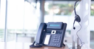 Ein Telefon steht auf einem Tisch in einer hellen Büroumgebung. Daneben hängt über eine Trennwand ein Headset.