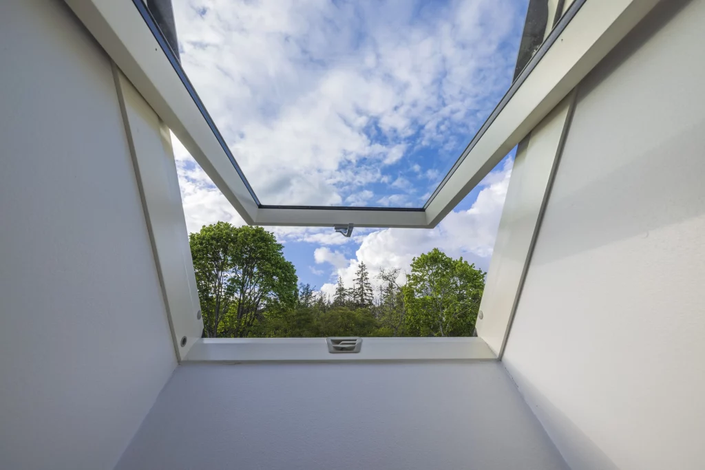 Geöffnetes Dachfenster mit Ausblick auf Bäume und einen teilweise bewölkten Himmel.