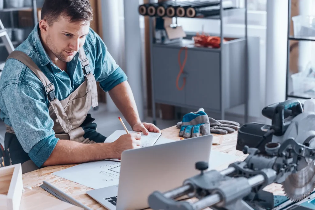 Handwerker in Arbeitskleidung sitzt an einer Werkbank mit Laptop, Bauplänen und Notizbuch. Er macht sich Notizen und informiert sich über Facotring im Handwerk, während eine Kreissäge und Arbeitshandschuhe im Vordergrund liegen.