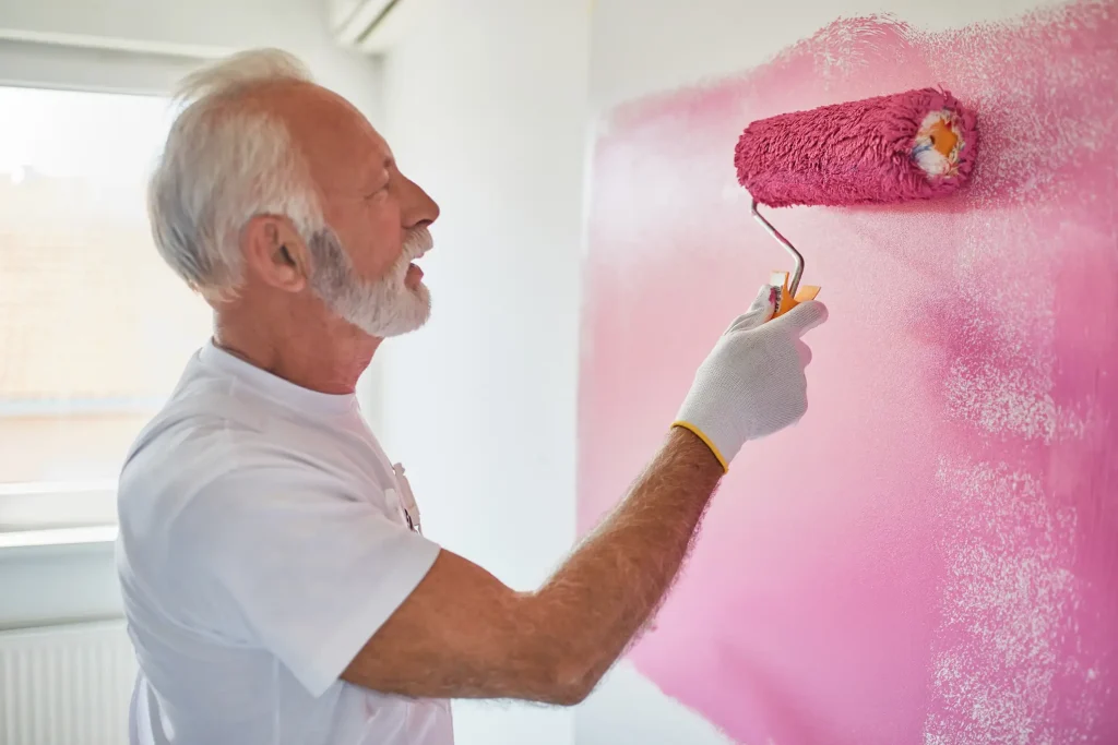 Ein älterer Maler mit grauem Bart streicht konzentriert eine Wand mit einer pinkfarbenen Farbrolle in einem hellen Raum.