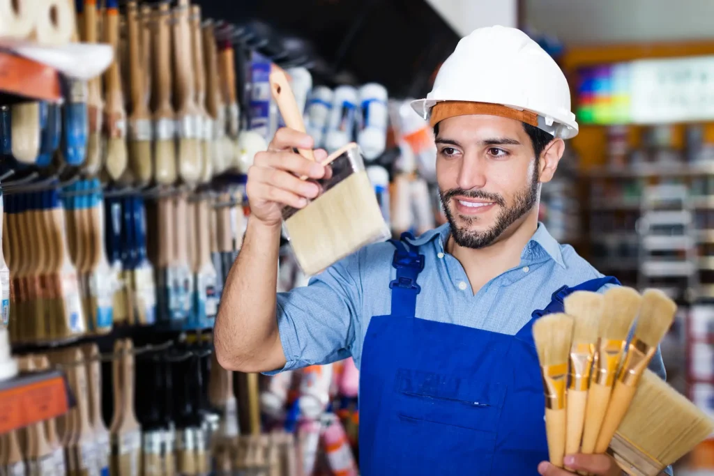 Ein Maler steht in seinem Farb- und Pinsellager und begutachtet einen Pinsel, während er mehrere weitere in der Hand hält.