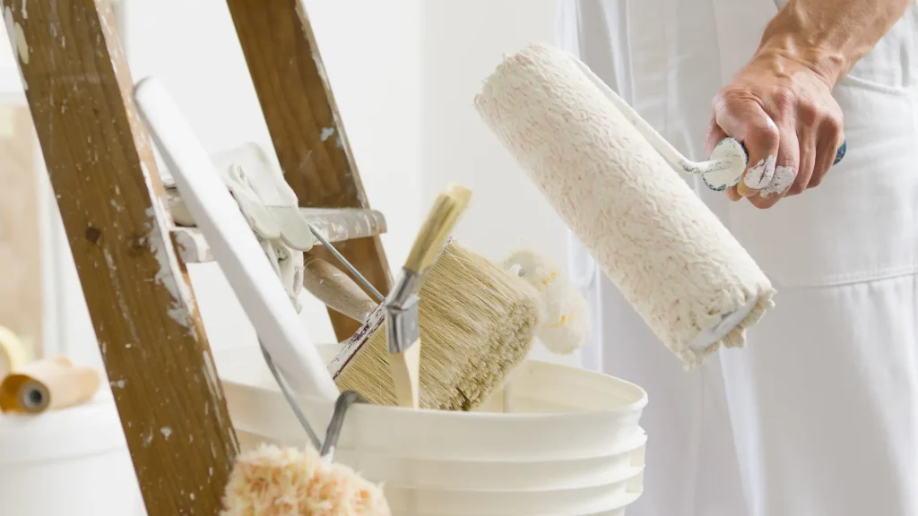 Ein Maler steht neben einer Holzleiter und bereitet seine Werkzeuge vor, in einem Eimer liegen Pinsel und eine Farbrolle, daneben liegen Arbeitshandschuhe.