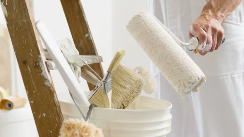 Ein Maler steht neben einer Holzleiter und bereitet seine Werkzeuge vor, in einem Eimer liegen Pinsel und eine Farbrolle, daneben liegen Arbeitshandschuhe.