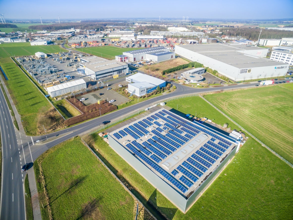 Luftaufnahme eines Industriegebiets mit mehreren Lager- und Produktionshallen, im Vordergrund ein Gebäude mit großflächiger Photovoltaikanlage auf dem Dach zur Nutzung von Solarenergie.