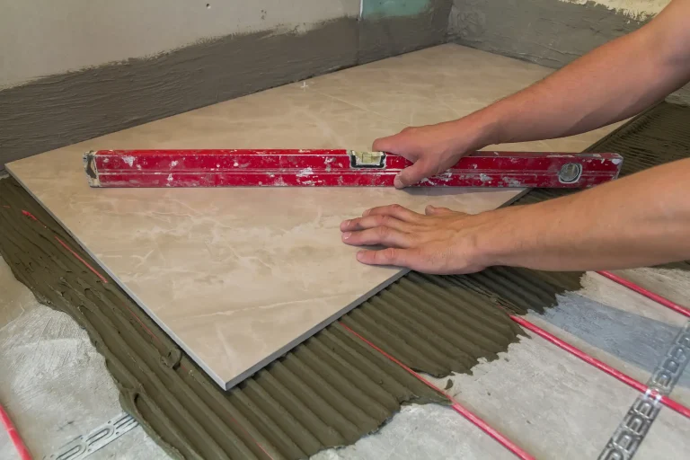 Ein Fliesenleger verlegt eine großformatige Keramikfliese auf einem grauen Kleberbett, unter dem die roten Rohre einer wasserführenden Fußbodenheizung sichtbar sind.