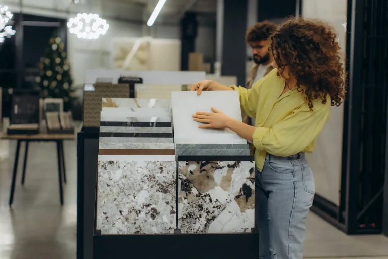 Eine Kundin in einem Fliesenfachhandel betrachtet verschiedene großformatige Fliesenmuster in einer Ausstellungshalterung. Sie begutachtet eine helle Fliese mit Marmor-Optik, während im Vordergrund weitere Naturstein-Designs zu sehen sind.